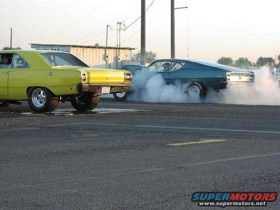 Yellow and teal muscle cars on a drag strip, one doing a burnout