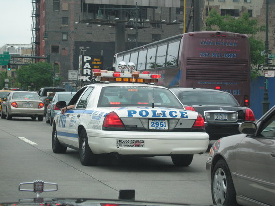 NYC police car with lights on, in traffic behind a bus and other cars