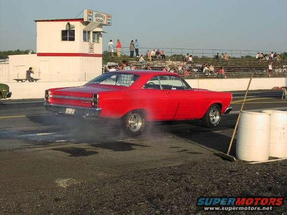 Red coupe with racing slicks, smoking tires, and a roll cage, viewed from the side at a drag strip