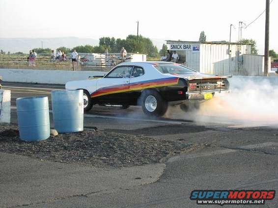 Drag racing car with rainbow stripe doing a burnout, kicking up smoke. Two blue barrels are in the foreground