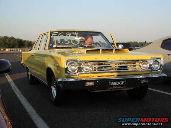 Yellow Plymouth Satellite with a hood scoop and a driver. Another car is parked next to it