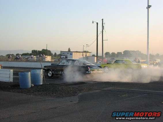 Black car and yellow car doing burnouts on a drag strip, kicking up smoke