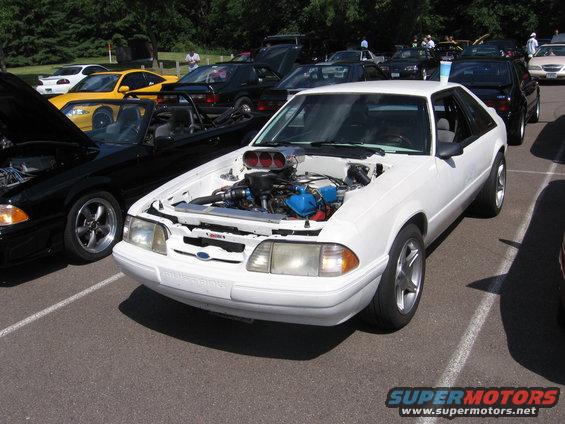 White Ford Mustang with hood open, showing a supercharger and blue engine components. Modified front end