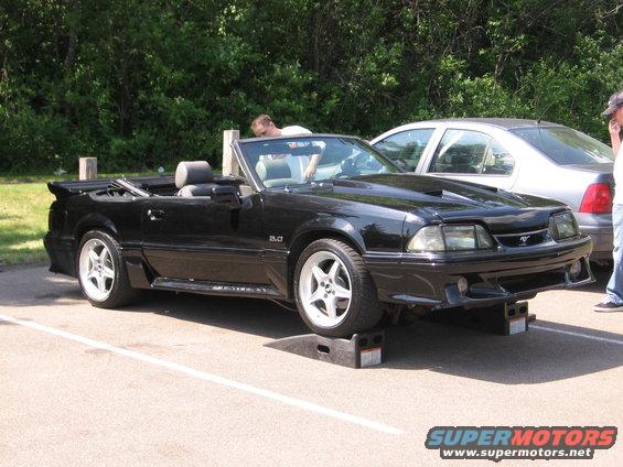 Black convertible Mustang on ramps, another car parked behind it