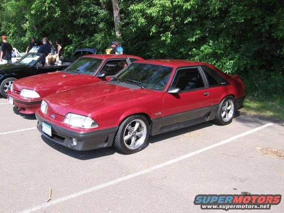 Red 1987 Ford Mustang coupe parked in a lot, showing its 3/4 front view with dark gray lower body cladding and aftermarket wheels