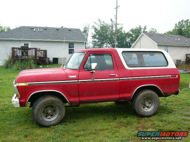 Side view of a red 1978 Ford Bronco with a white top parked on grass in front of houses