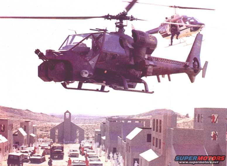 Two helicopters fly over a model town with a church and cars