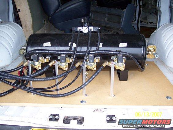 Interior of a van showing a black air suspension tank with multiple solenoid valves and hoses mounted on a wooden platform