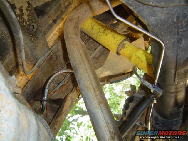 Close-up of vehicle undercarriage showing exhaust pipe, brake lines, and yellow shock absorber. Supermotors.net logo visible