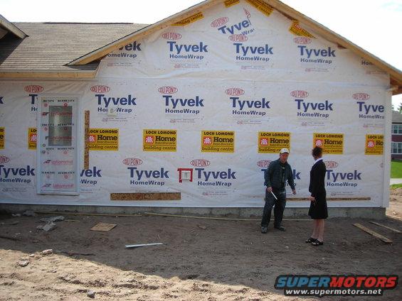 Construction site with house framing covered in Tyvek wrap and two people standing on dirt