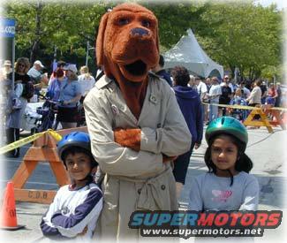 Person in a large dog costume stands with arms crossed between two children wearing helmets. Crowd in background