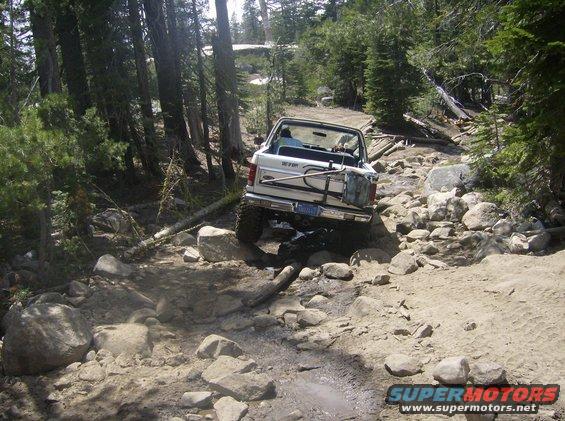 White SUV navigating a difficult, rocky, wooded trail. Spare tire mounted on the back