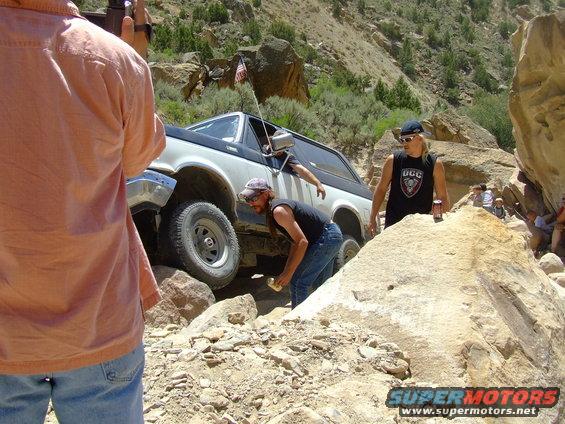 White and black Ford Bronco navigating a rocky trail, driver's side front wheel high, with people watching