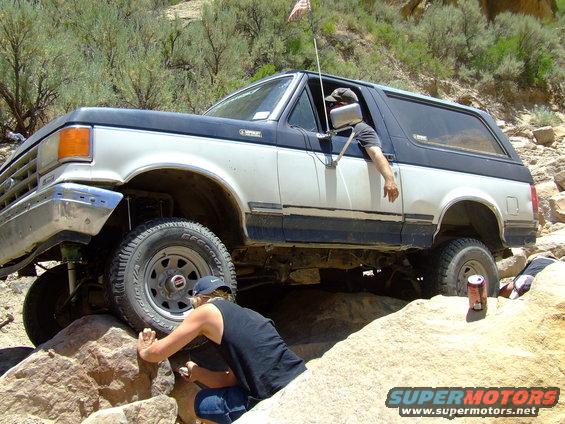 White and blue 1987 Ford Bronco off-roading over rocks, driver's arm out window