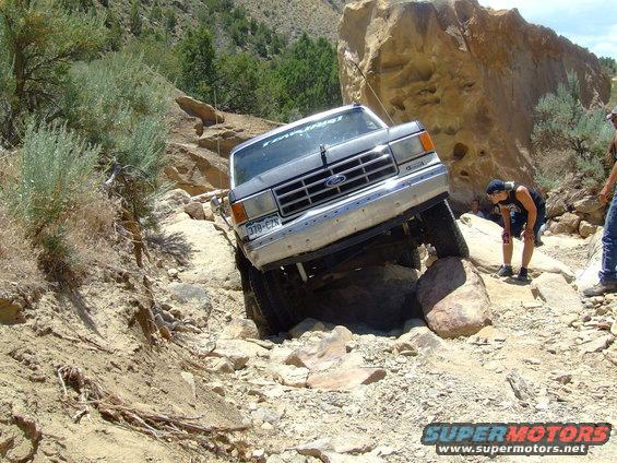 Silver 1987 Ford Bronco navigating a rocky, off-road trail with its front passenger side tire high on a boulder