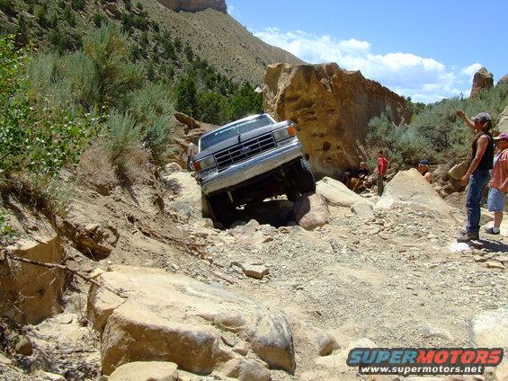 Silver 1987 Ford Bronco navigating a rocky off-road trail with its front wheels on large boulders