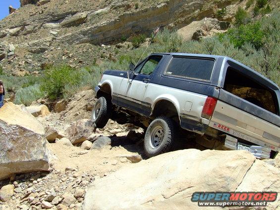 Two-tone 1987 Ford Bronco navigating a rocky off-road trail, with its front passenger side wheel lifted