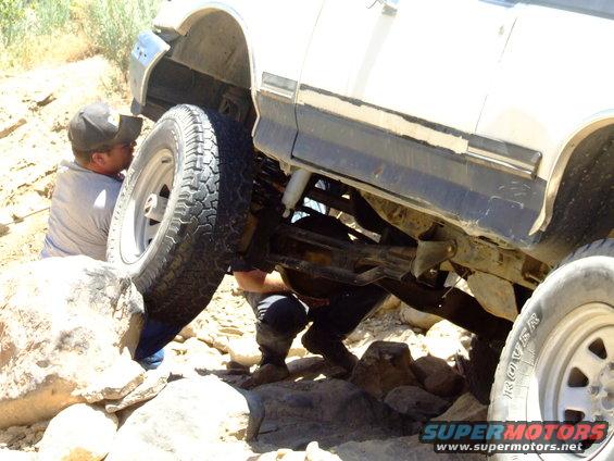 1987 Ford Bronco with front wheel high on rock, people working underneath