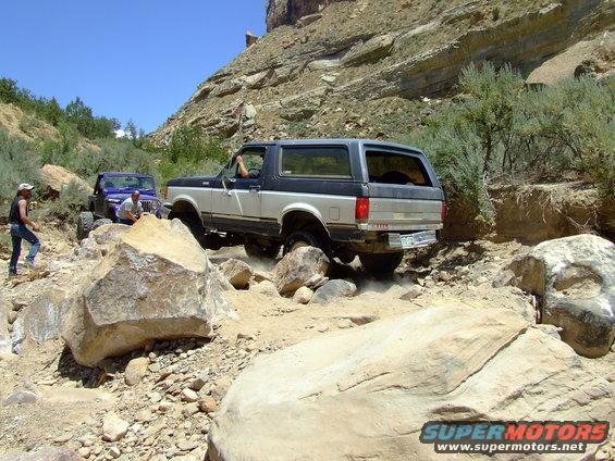 1987 Ford Bronco rear view, two-tone silver and blue, navigating a rocky off-road trail with another Jeep behind it
