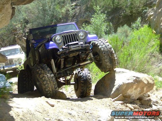 Purple Jeep Wrangler climbing over a large rock on a dirt trail, with a white SUV behind it