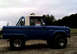 1973 Ford Bronco - A blue 1973 Ford Bronco with a white roof and roll bar is parked on a dirt lot under a cloudy sky.