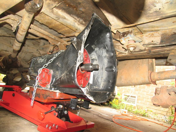 Transmission being lowered by a red jack, with driveshaft and exhaust visible