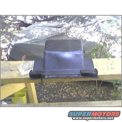 Black plastic automotive part with rectangular openings on the sides, resting on a wooden surface