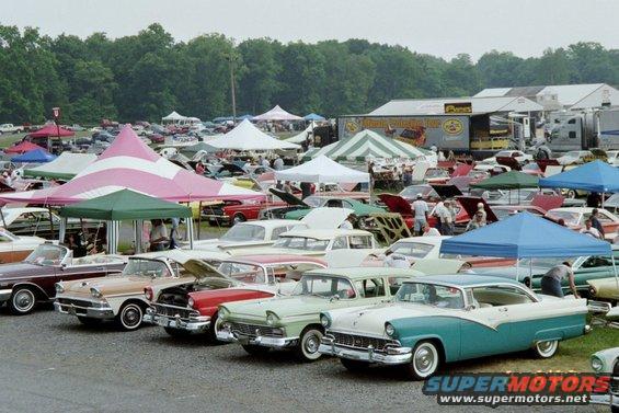 Many classic cars parked under tents at an outdoor event. A turquoise and white 1956 Ford Fairlane is in the foreground