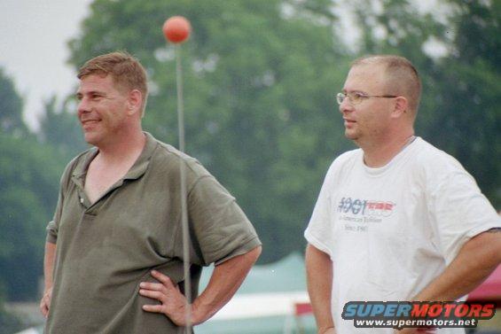 Two men stand outdoors, one in a green polo, the other in a white t-shirt with text
