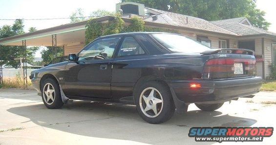 Black 1989 Mazda MX-6 coupe parked on a concrete driveway, rear quarter view. The car has a spoiler and aftermarket wheels