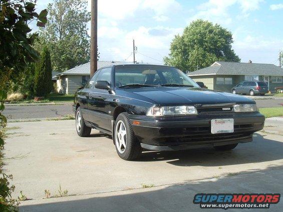 Black 1989 Mazda MX-6 coupe parked on a concrete driveway, front 3/4 view. The car has a license plate and a GT badge