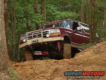 1995 Ford Bronco with custom bumper and lift kit driving on a dirt trail in a forest