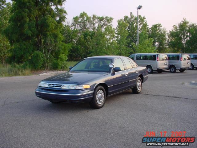 Dark blue 2003 Ford Crown Victoria police car parked on asphalt, with silver vans in the background