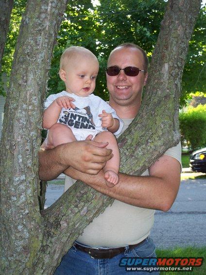 Man holding a baby while standing between two tree trunks. A car is visible in the background