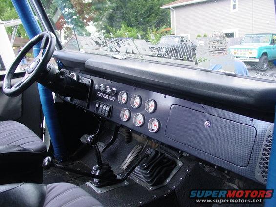 Interior of a 1973 Ford Bronco showing dashboard, steering wheel, gauges, and gear shifter. Two other Broncos are visible outside