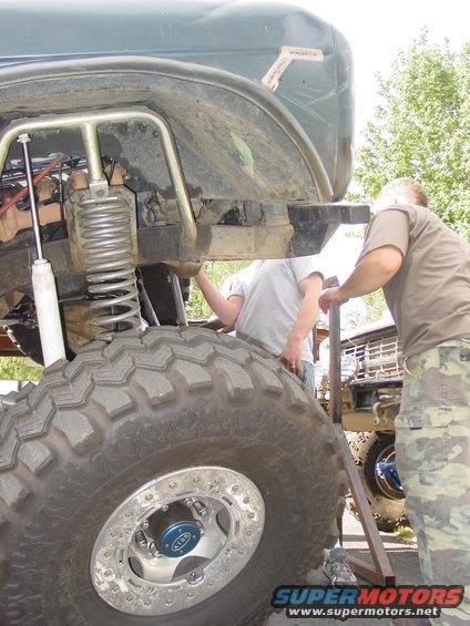 Close-up of a large off-road tire and suspension components with two people working on the vehicle