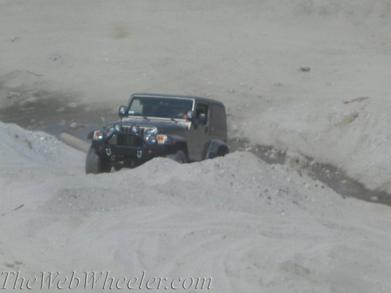 Dark-colored Jeep Wrangler navigating a sandy, uneven terrain with auxiliary lights on the front bumper
