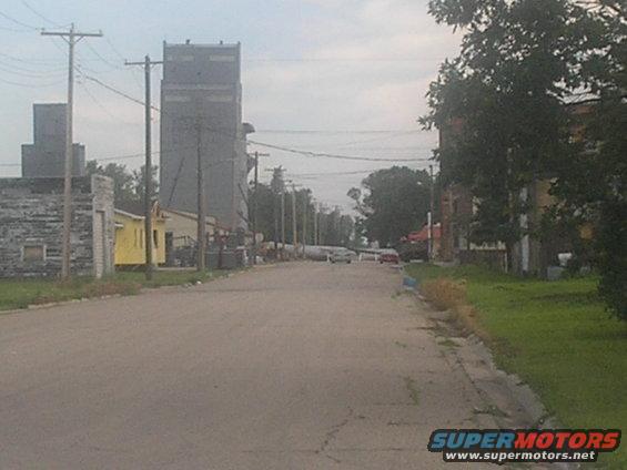 Street view with tall industrial buildings, a yellow building, and cars on the road. Supermotors logo in corner