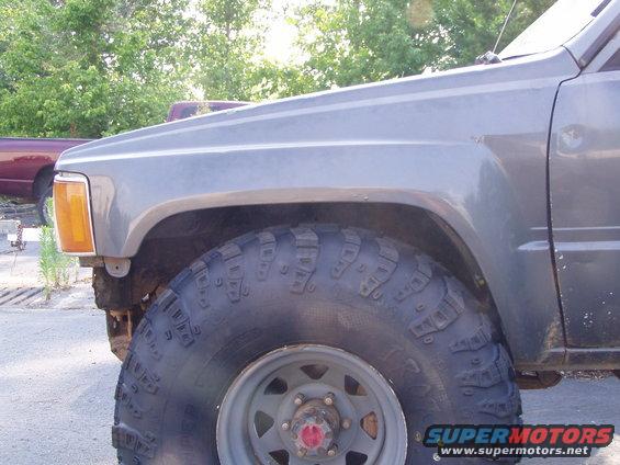 Gray truck fender and large off-road tire with red hub cap. Another maroon truck is visible in the background