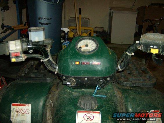 Green ATV dashboard with gauges, ignition, and handlebars. Warning labels are visible on the body
