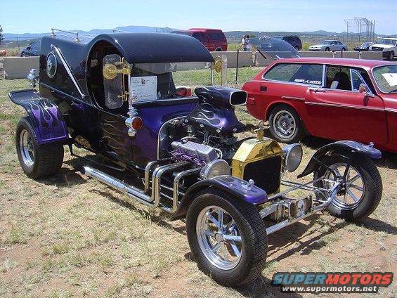 Custom hot rod with purple flames, exposed engine, and chrome exhaust pipes parked next to a red station wagon