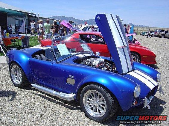 Blue AC Cobra replica with hood open, parked at an outdoor car show. Red car behind it
