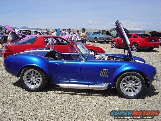 Blue convertible sports car with side pipes and hood open, parked on gravel