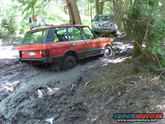 Red Range Rover stuck in mud, another SUV behind it on a wooded trail