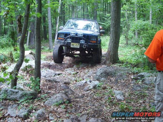 Blue Jeep Cherokee XJ with aftermarket bumper and large tires on rocky terrain