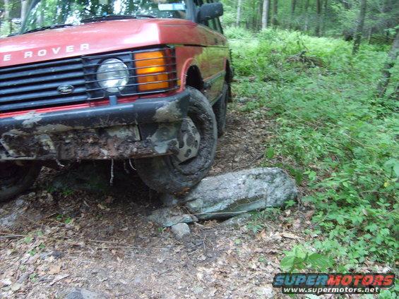 Red Range Rover driving over a rock on a dirt path in a wooded area