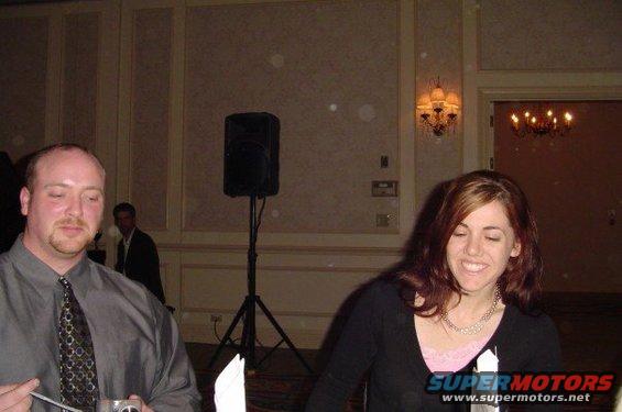 Man in a grey shirt and patterned tie, woman in a black top, indoor event with speakers and chandeliers