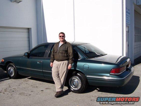 Man in jacket and khakis stands by a dark green sedan. The car has chrome hubcaps and a chrome strip along the bottom