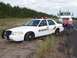 2003 Ford Crown Victoria - A white 2003 Ford Crown Victoria sheriff's car is stuck in a dirt ditch beside a highway, with a tow truck and a man attempting to assist.