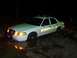 2003 Ford Crown Victoria - A white sheriff's car is stuck in a muddy area at night, with its lights on.